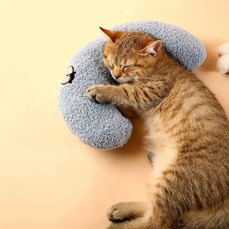 A brown cat sleeps curled around a gray crescent-shaped toy on a beige surface, snuggled against The Stuff Box Cozy Pet Pillow for Cats and Small Dogs made of soft and cozy material.