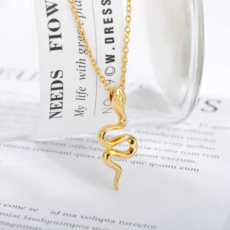 The Snake Pendant, a stylish necklace featuring a gold pendant in the form of a snake, hangs from a gold chain, draped over a glass container placed on a paper containing scattered text.