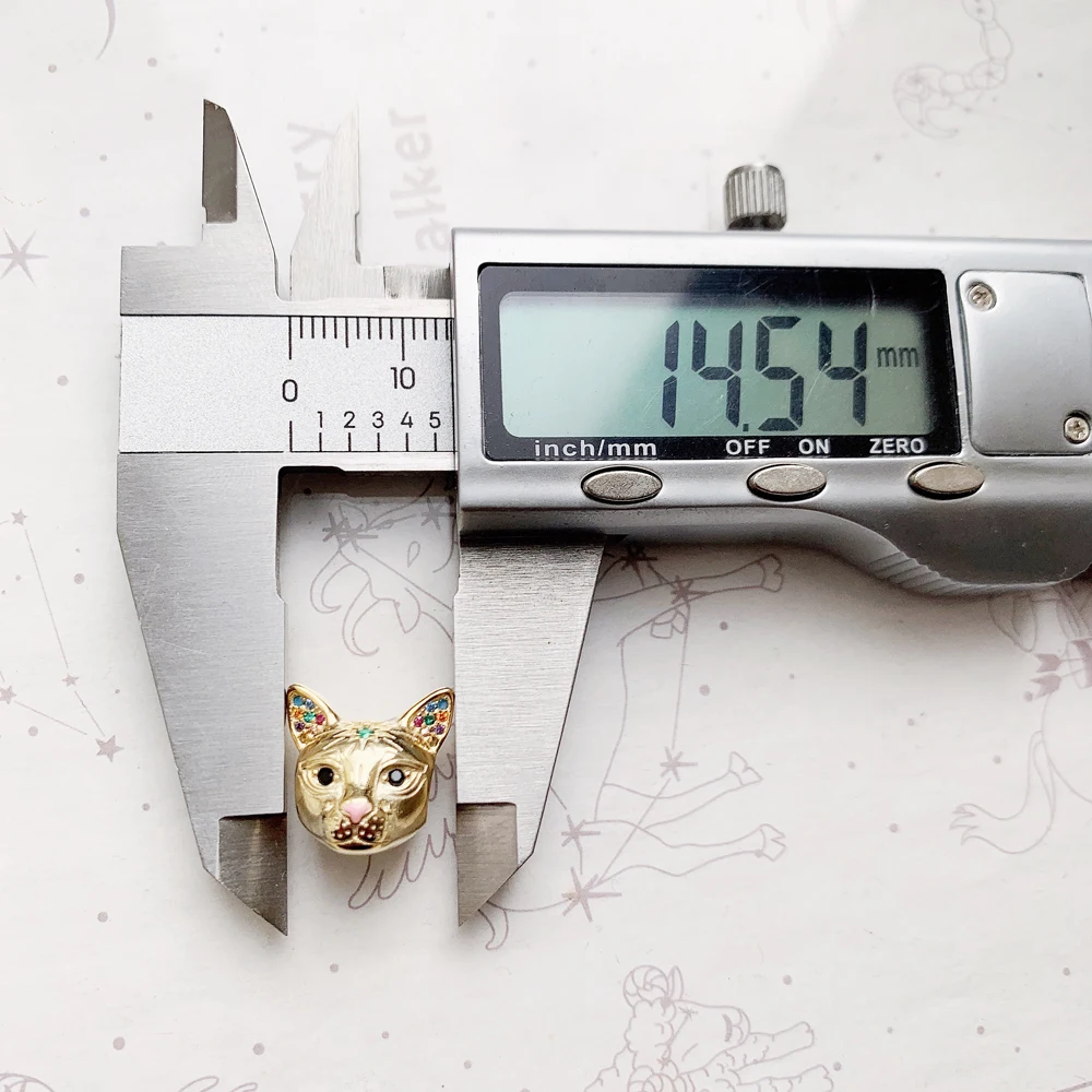 A luxury gold plated cat head charm bead with an emerald green forehead stone and pink enamel nose showing dimensions on a digital caliper