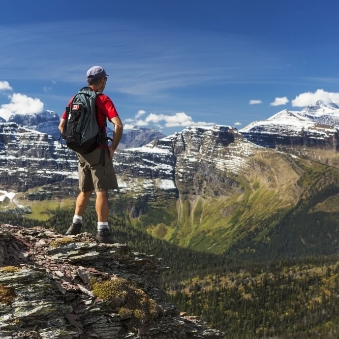 Male hiker standing on top of mountain ridge overlooking snow peaked mountains and forest valley below with blue sky | Дом и сад