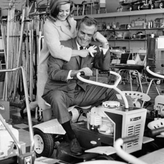 Mid adult man sitting on a lawn mower with a mid adult woman standing behind him in a store Poster Print (24 x 36)