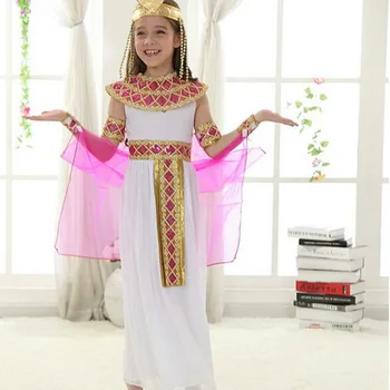 Petite fille portant le déguisement Cléopâtre pour fille se composant d'une longue robe blanche et fluide avec une ceinture rose et dorée, un large plastron doré et rose couvrant les épaules, une couronne avec des mèches pendantes dorées, des bracelets et une toge rose, mis en scène à l'interieur d'une maison.