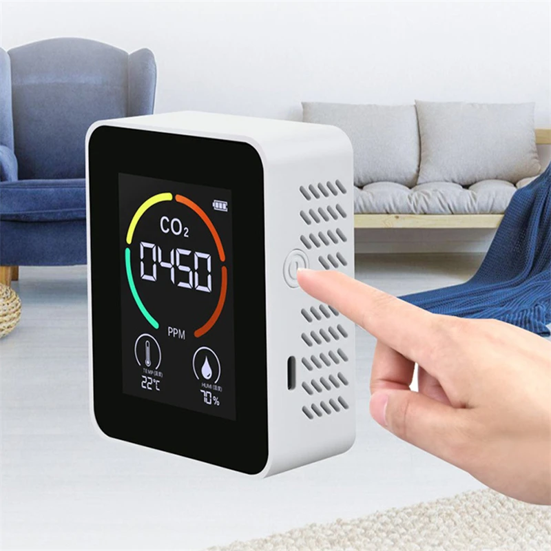 A person's hand presses a button on a Carbon Dioxide Detector displaying 450 PPM with indicators for temperature and humidity, placed in a living room with a sofa and chair in the background, ensuring optimal air quality control.
