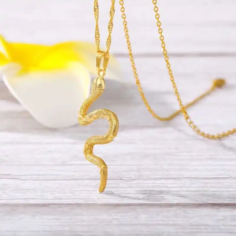 The Snake Pendant, a chic addition to women's fashion accessories, delicately hangs on a chain displayed on a wooden surface with a yellow and white flower in the background.