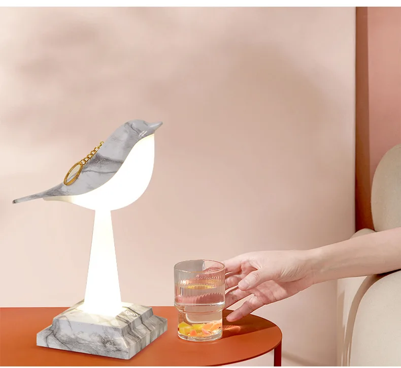 A hand reaches for a clear glass of water placed on a red surface beside the Bird Table Night Light, an exquisite piece of contemporary design with a marble base and elegant craftsmanship, complete with a bracelet hanging gracefully from its beak.