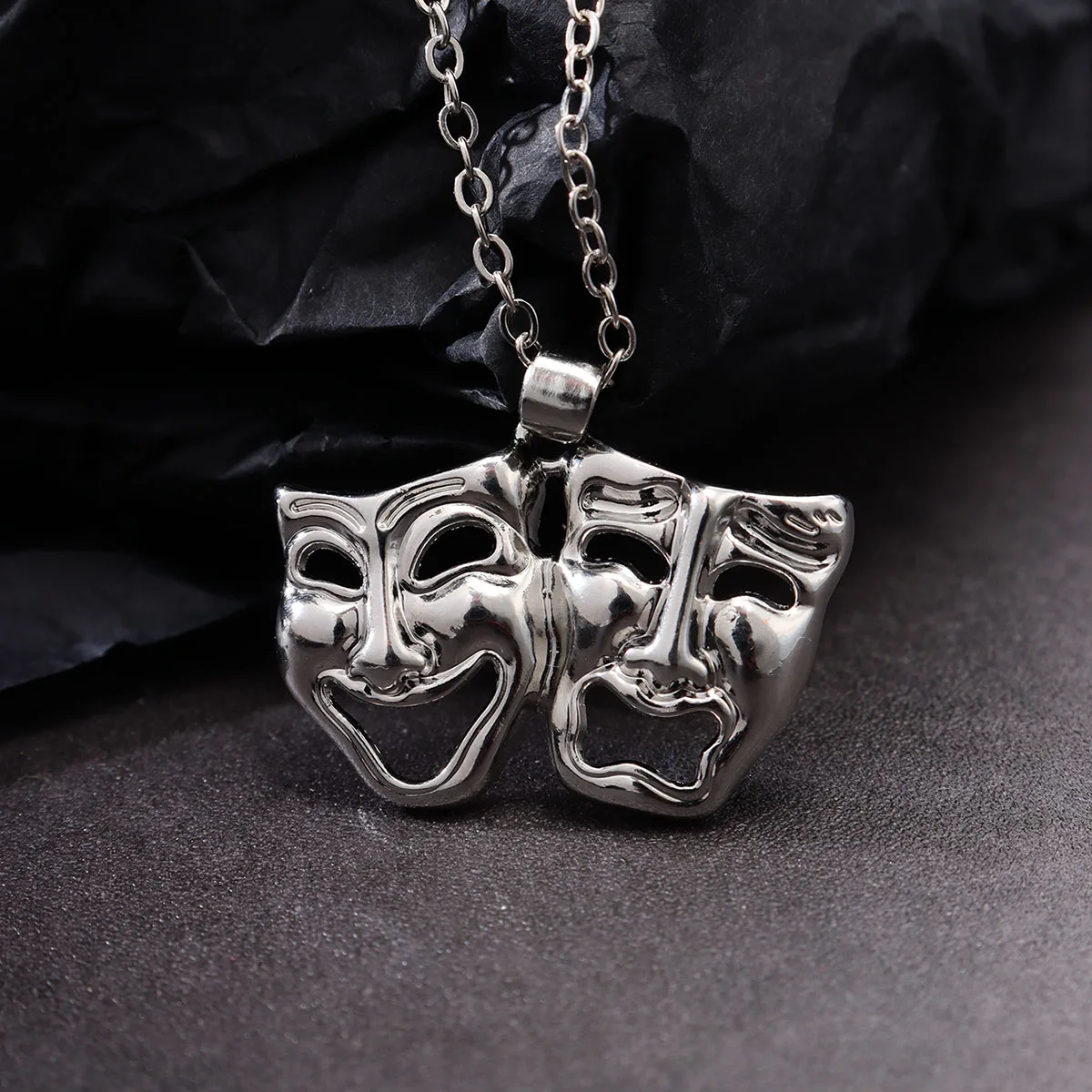 Masque de théâtre argenté en métal, pendentif gothique tendance sur fond noir, reflets lumineux.
