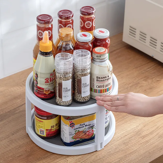 A white rotating storage tray on a kitchen counter, holding various spice jars and bottles, with a hand spinning it.