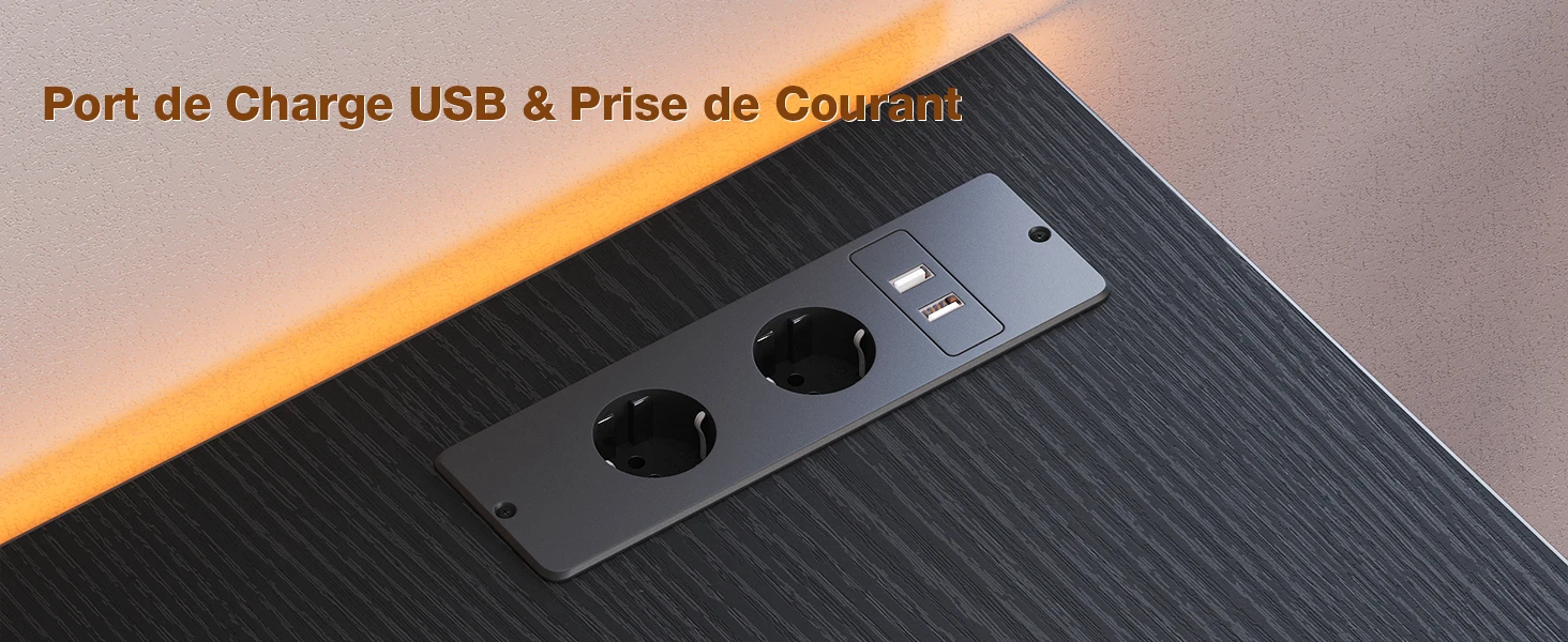 Close-up of a desk edge with a built-in USB charging port and power outlet. Dark wood finish with orange mood lighting above.