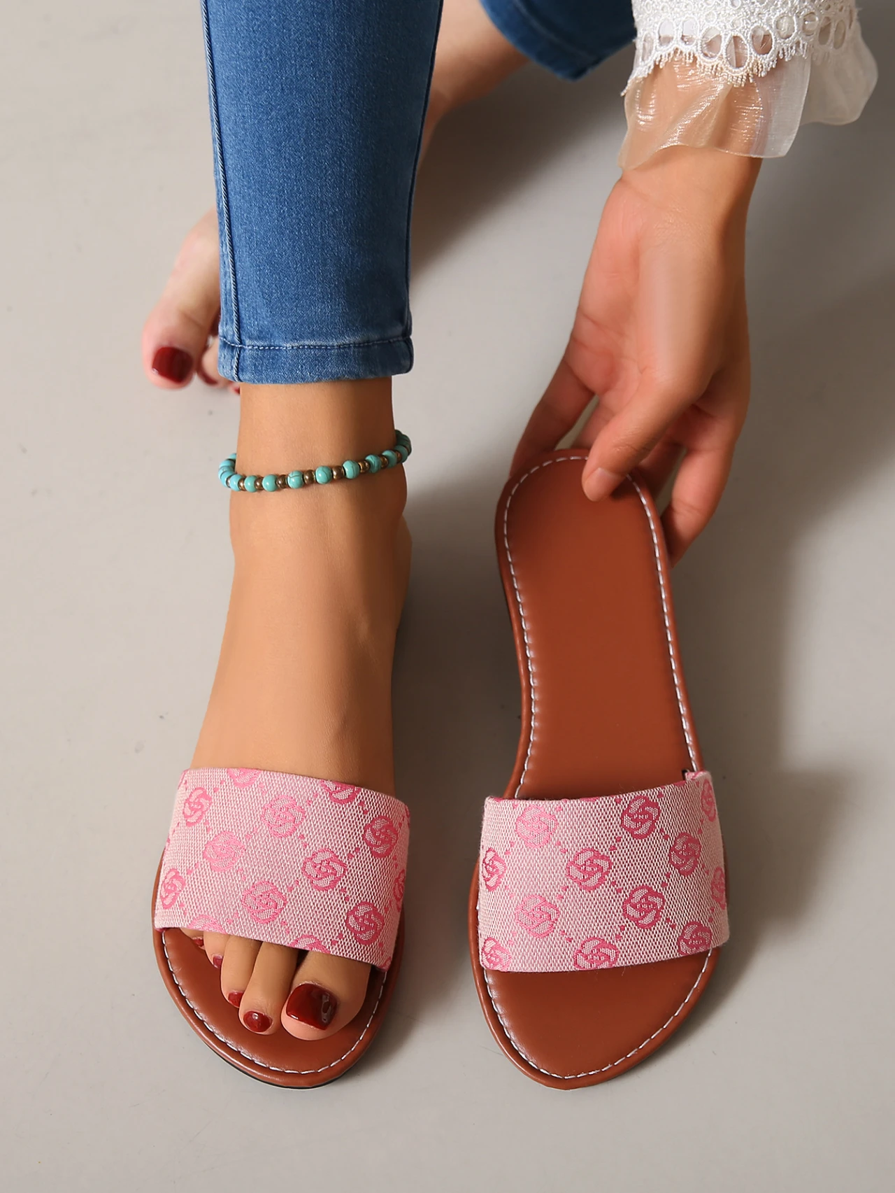 Wearing blue jeans, a white lace sleeve, and a turquoise anklet, someone tries on French Sole pink open-toe sandals with a patterned strap—modern British-inspired slippers ideal for summer, highlighting red-painted toenails.