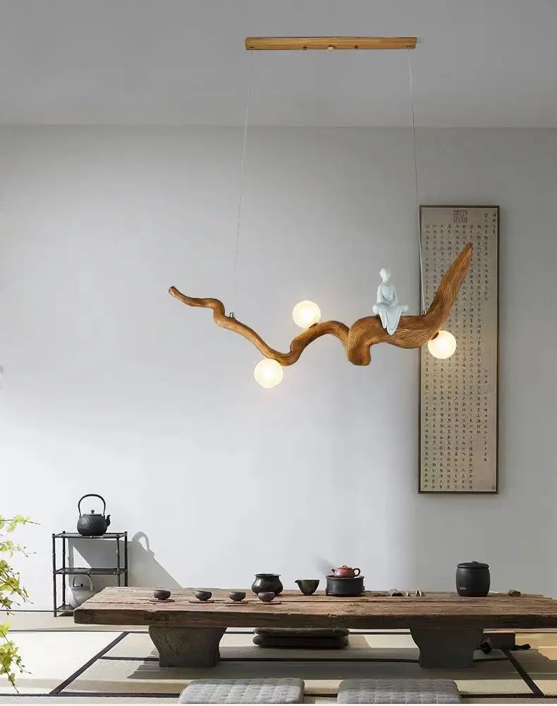 A modern room showcases a long wooden table and cushions. Above, a Zen-Inspired Wooden Pendant Light resembling a branch with three glowing bulbs and a small figurine enhances the zen ambiance. Minimalist decor and a tea set complete the serene aesthetic.