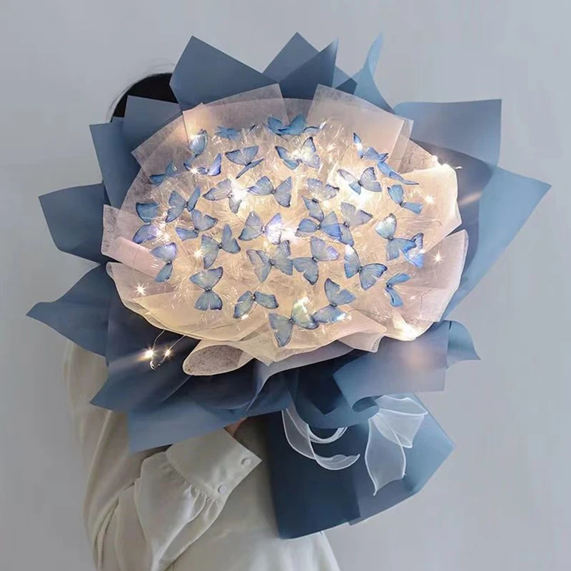 A person holds a Creative Handmade Butterfly Bouquet featuring light blue and white paper butterflies illuminated by glowing lights, wrapped in blue and white translucent paper. This exquisite piece, known as the Flower Material Package for DIY Bride Wedding Decor, functions as both a stunning home decoration and an ethereal display of artificial flowers, making it ideal for romantic Valentine's Day gifts.