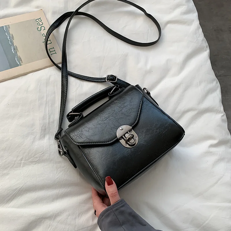 Ehtne must nahast väike crossbody kott sangaga, musta rihmaga, valgel voodil lebamas. Koti eesosas on metallist lukk.