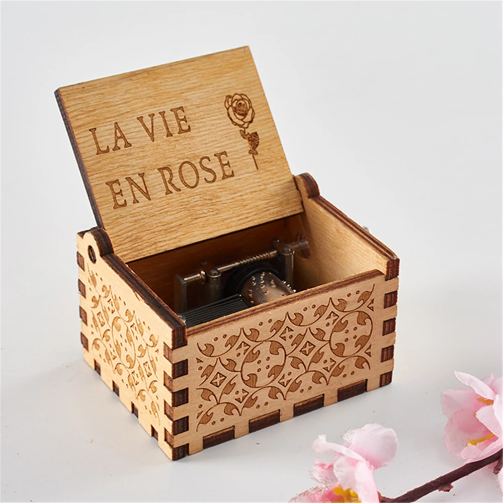 An open Wooden Music Box with intricate engravings on the sides. The lid is adorned with the words "LA VIE EN ROSE" and a rose design. A small gear mechanism and a pink flower are visible nearby, capturing the charm of classic tunes in this handmade music box.