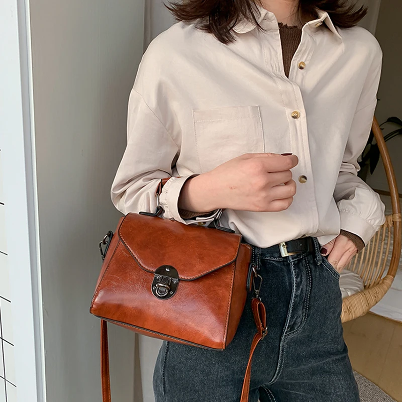 Naine hoiab punakaspruuni väikest crossbody kotti sangaga, millel on metallist lukk. Kott on õlakoti stiilis ja sobib igapäevaseks kandmiseks. Taustal on hele ruum ja valge sein.