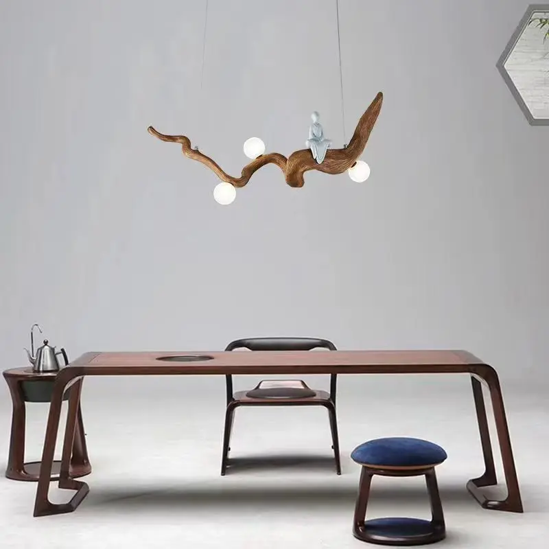 A minimalist room showcases a wooden table, chair, and stool. The Zen-Inspired Wooden Pendant Light, handcrafted to resemble branches with small globes, beautifully lights up the space. A hexagonal wall decor piece adds depth to the serene atmosphere.