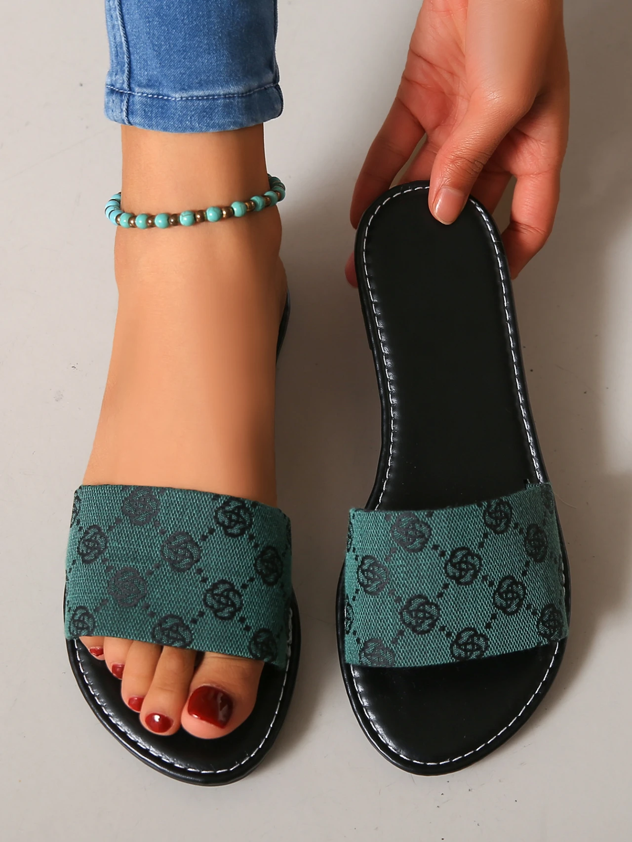 Wearing blue jeans and a turquoise beaded anklet, someone tries on French Sole’s green and black patterned modern slippers with black soles—one on, one in hand—ideal for adding British style to your summer outfit.