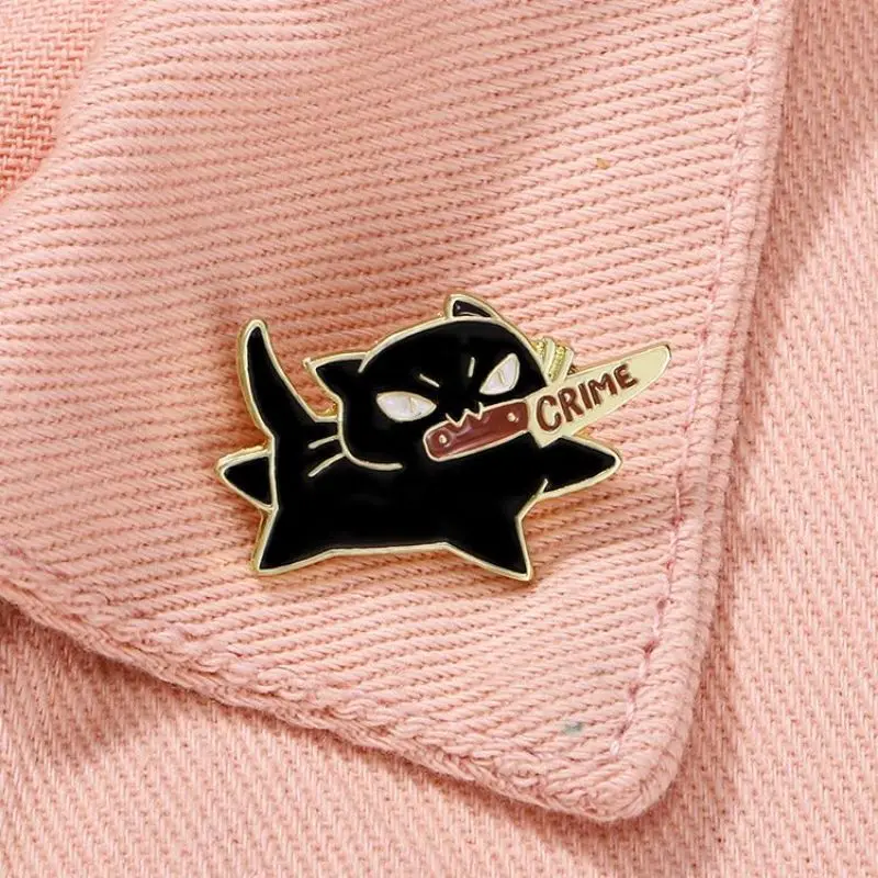 The Cute Black Cat Enamel Brooch by KittyNook Cat Co. holds a "CRIME" knife on a pink textured fabric background.