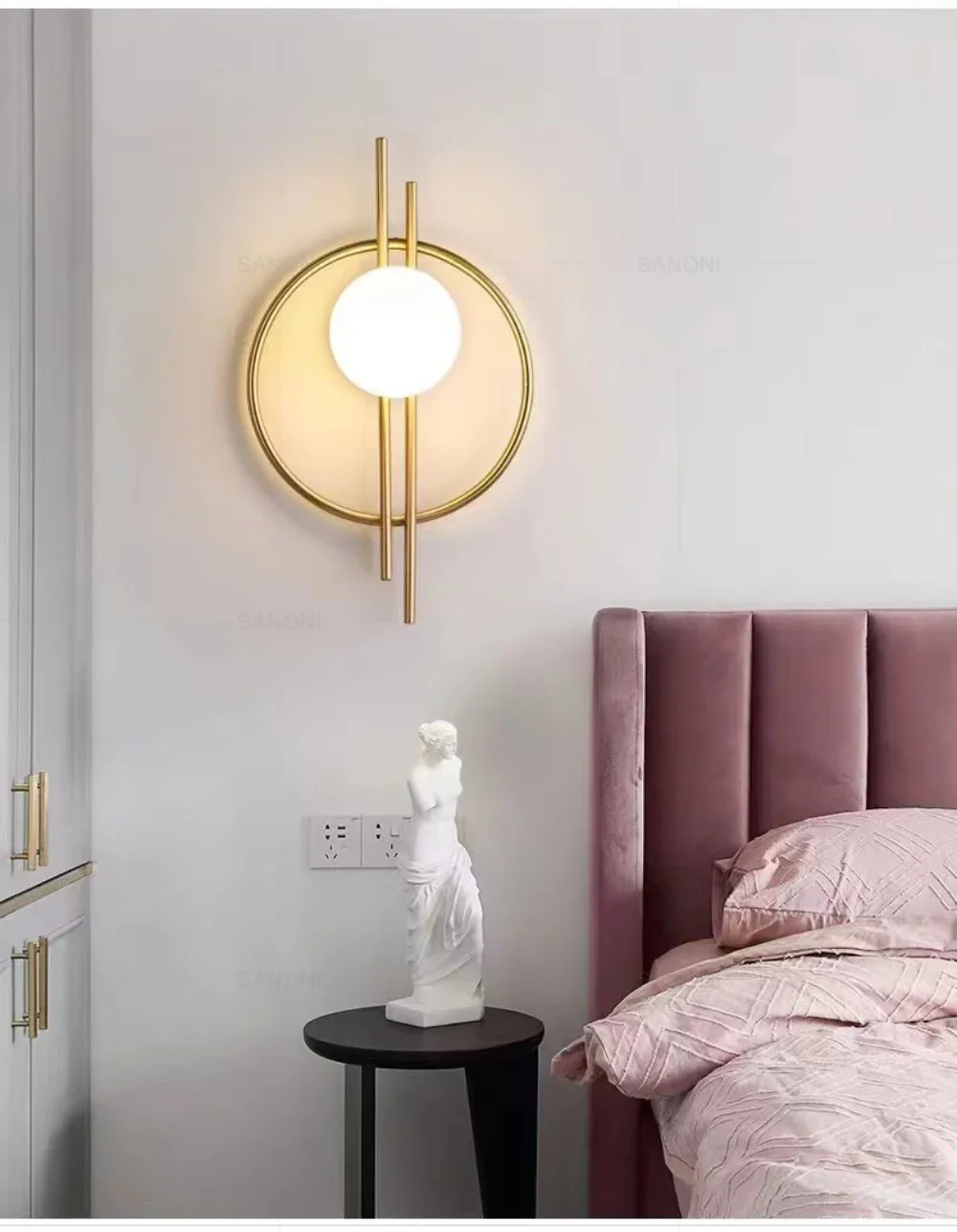 A modern bedroom featuring a pink upholstered headboard, a Circular Wall Sconce with Dual Vertical Rods, and a small side table holding a white statue.