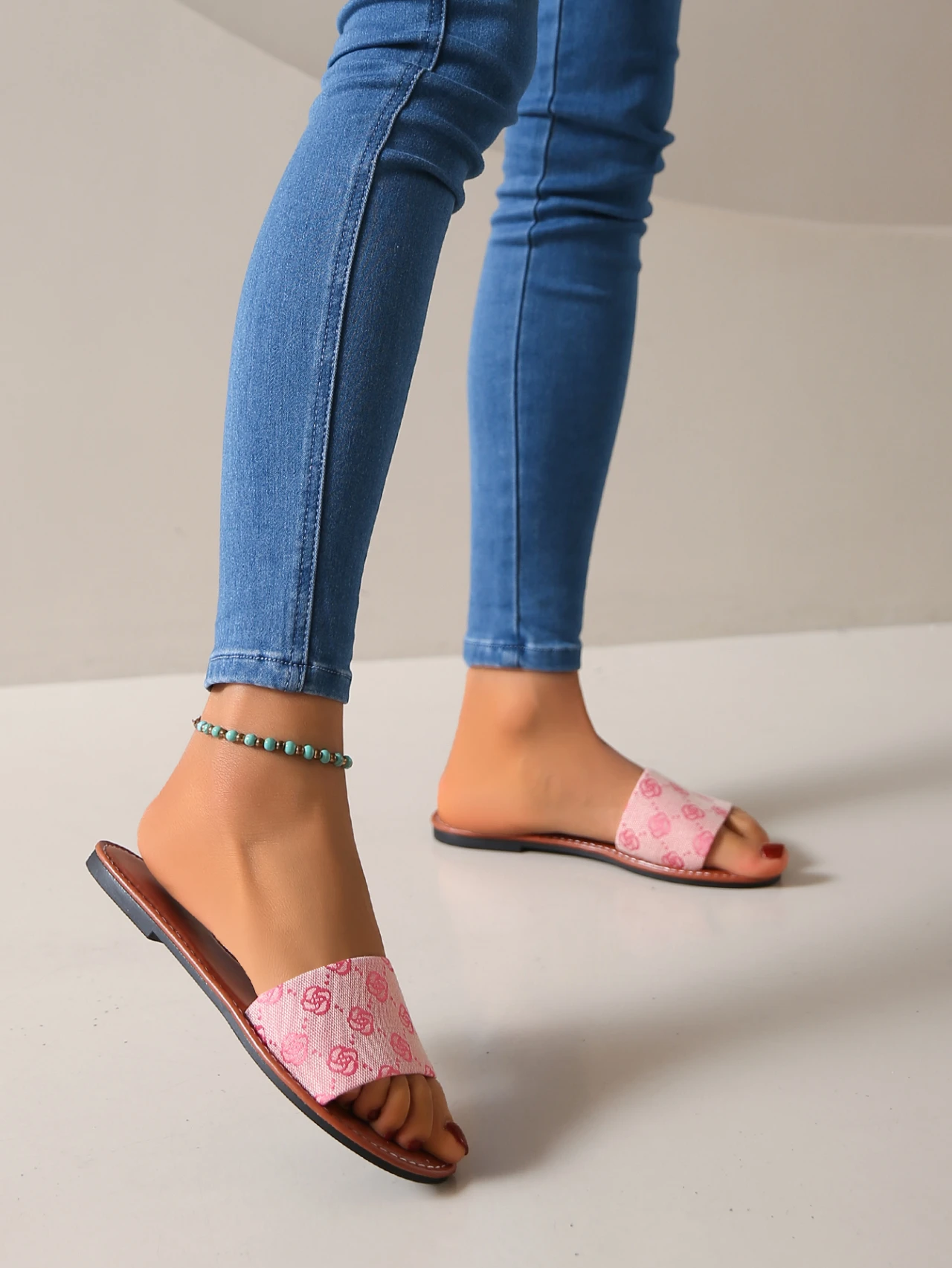 Wearing blue skinny jeans, a turquoise beaded anklet, and French Sole pink patterned modern slippers with brown soles, a person stands on a light floor, their footwear and accessories adding playful summer flair.