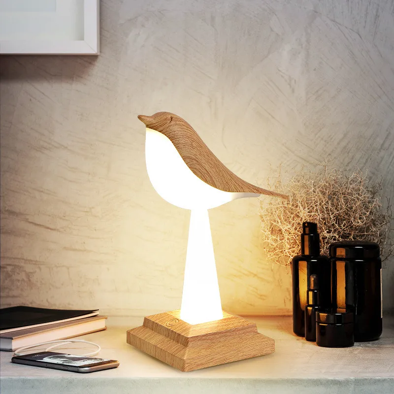 A Bird Table Night Light with elegant craftsmanship glows on a desk next to black bottles, a stack of books, and eyeglasses, epitomizing contemporary design.