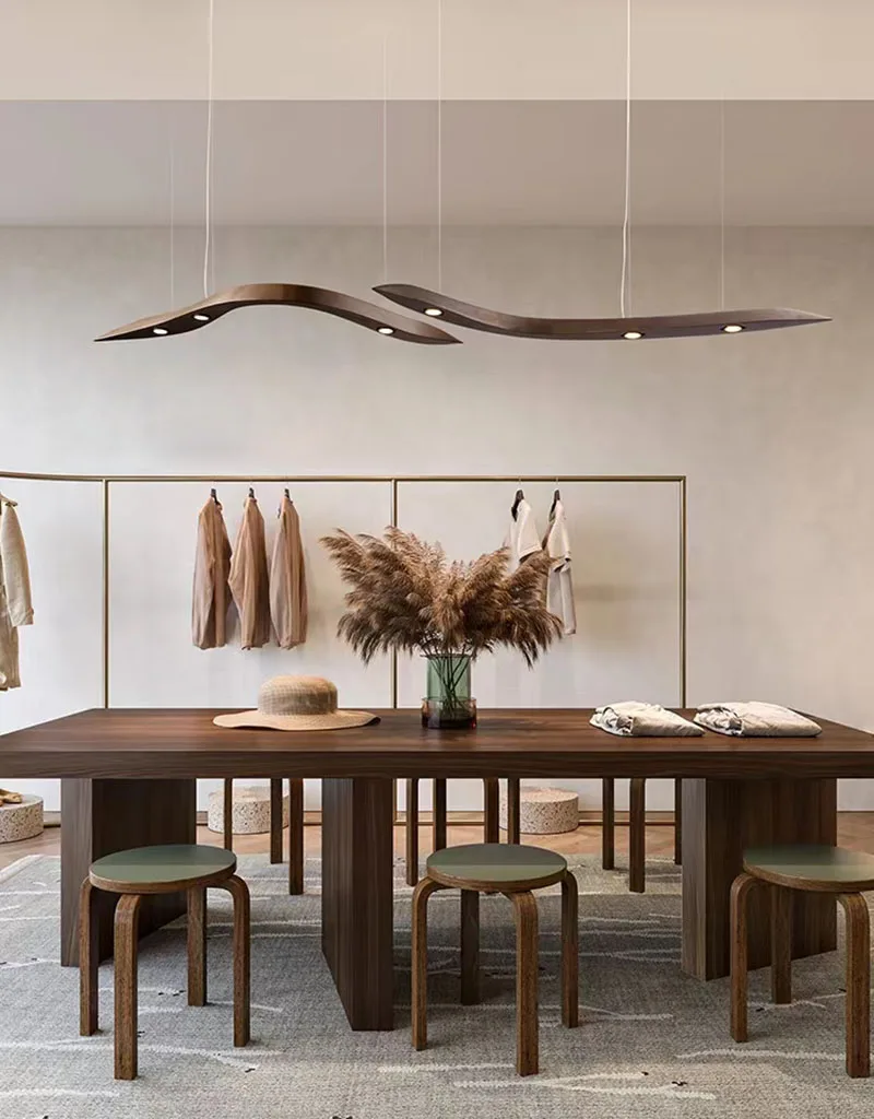 A minimalist room features a wooden table, four stools, a glass vase with pampas grass, hanging clothes, and the Spiral Wood Pendant Light, which brings an organic modern lighting touch to the space.