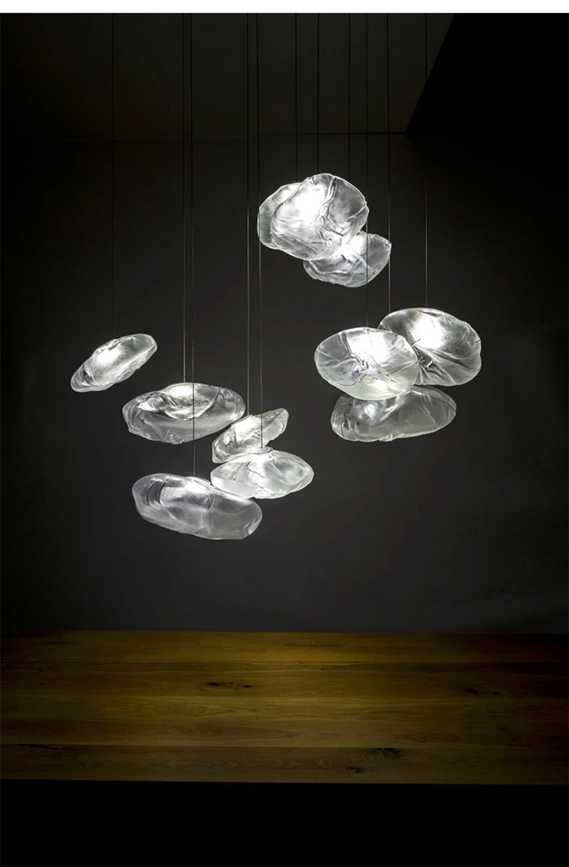 The Luxe Cloud Glass Pendant Light, cloud-like and translucent, is suspended from the ceiling and illuminated against a dark background above a wooden surface.