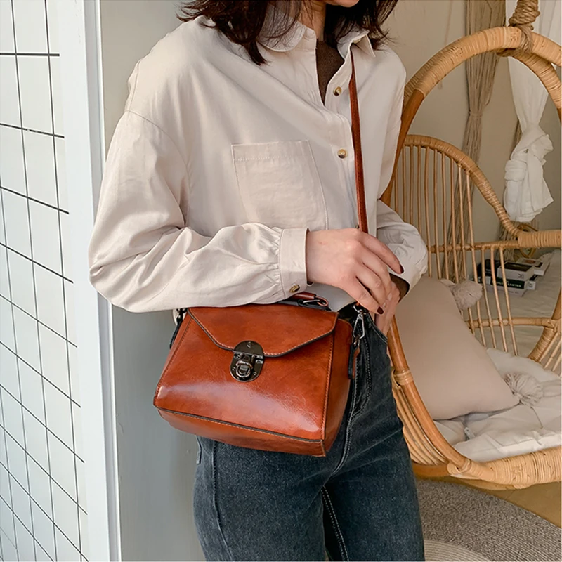 Naine kannab pruunist nahast väikest crossbody kotti sangaga, seistes modernse interjööriga ruumis. Koti lukustatav esikülg ja mugav õlarihm loovad stiilse ja praktilise ilme.