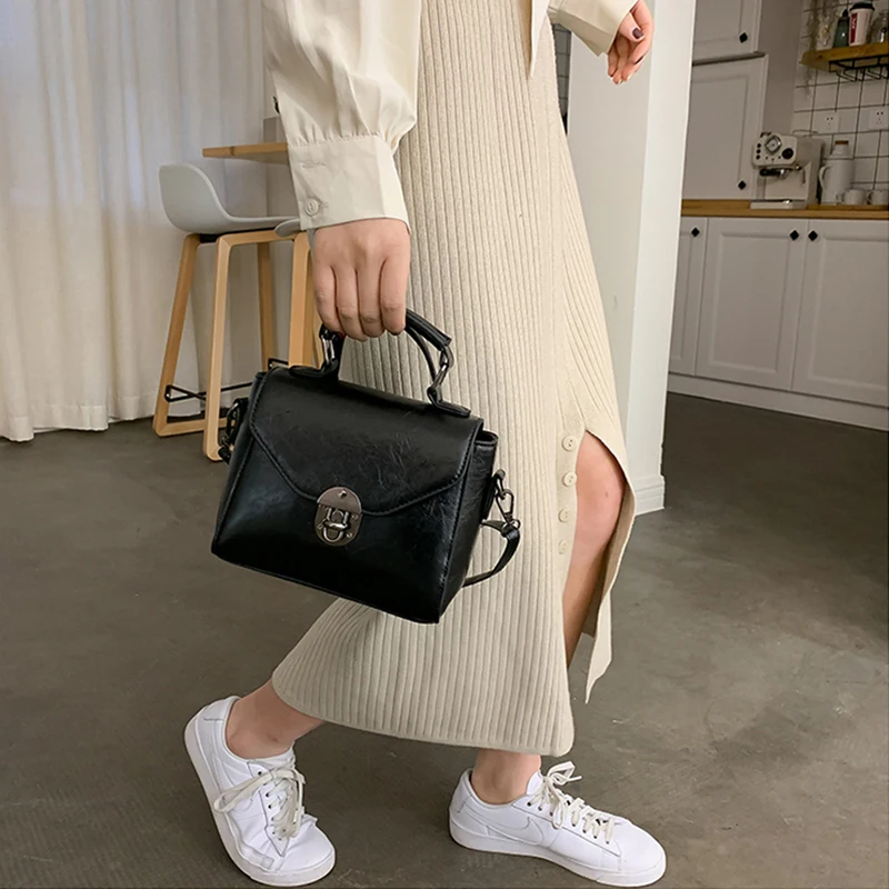 Inimene kannab väikest musta crossbody kotti sangaga, riietatud heledasse kudumisse ja valgetesse tossudesse, taustal kaasaegne köök.