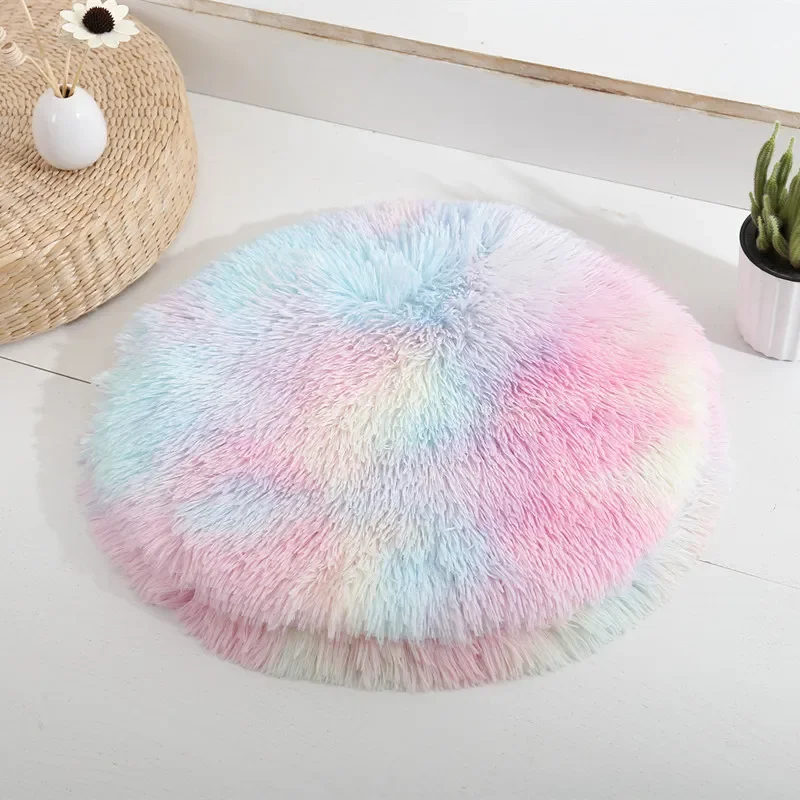 The Plush Cat Bed Pet Sleeping Mat Round Cat Cushion, featuring a soft pastel gradient of pink, blue, and yellow, rests upon a white surface. In the background, a small potted plant and a woven mat with a vase add to the cozy nook vibe.