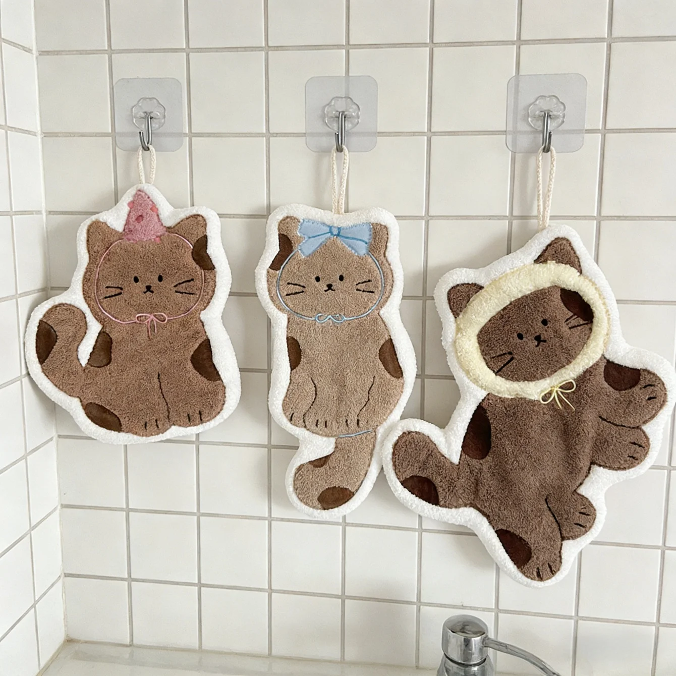 Three Soft Cat Paw Hand Towels by KittyNook Cat Co. hang above a sink, each with unique accessories and brown details.