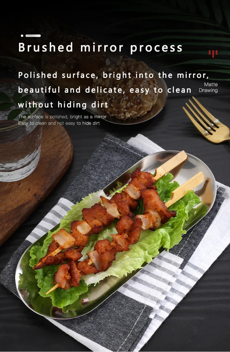An oval mirror-finish metal tray holding three grilled meat skewers placed over fresh lettuce leaves. The tray rests on a checkered napkin atop a black table, with a glass of water, a gold fork, and a wooden board visible in the background.
