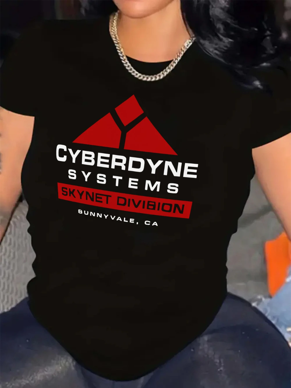Das schwarze T-Shirt zeigt eine rot-weiße Cybernet-Systemgrafik mit einem dreieckigen Design und dem Wort „SKYNET DIVISION“ darauf.