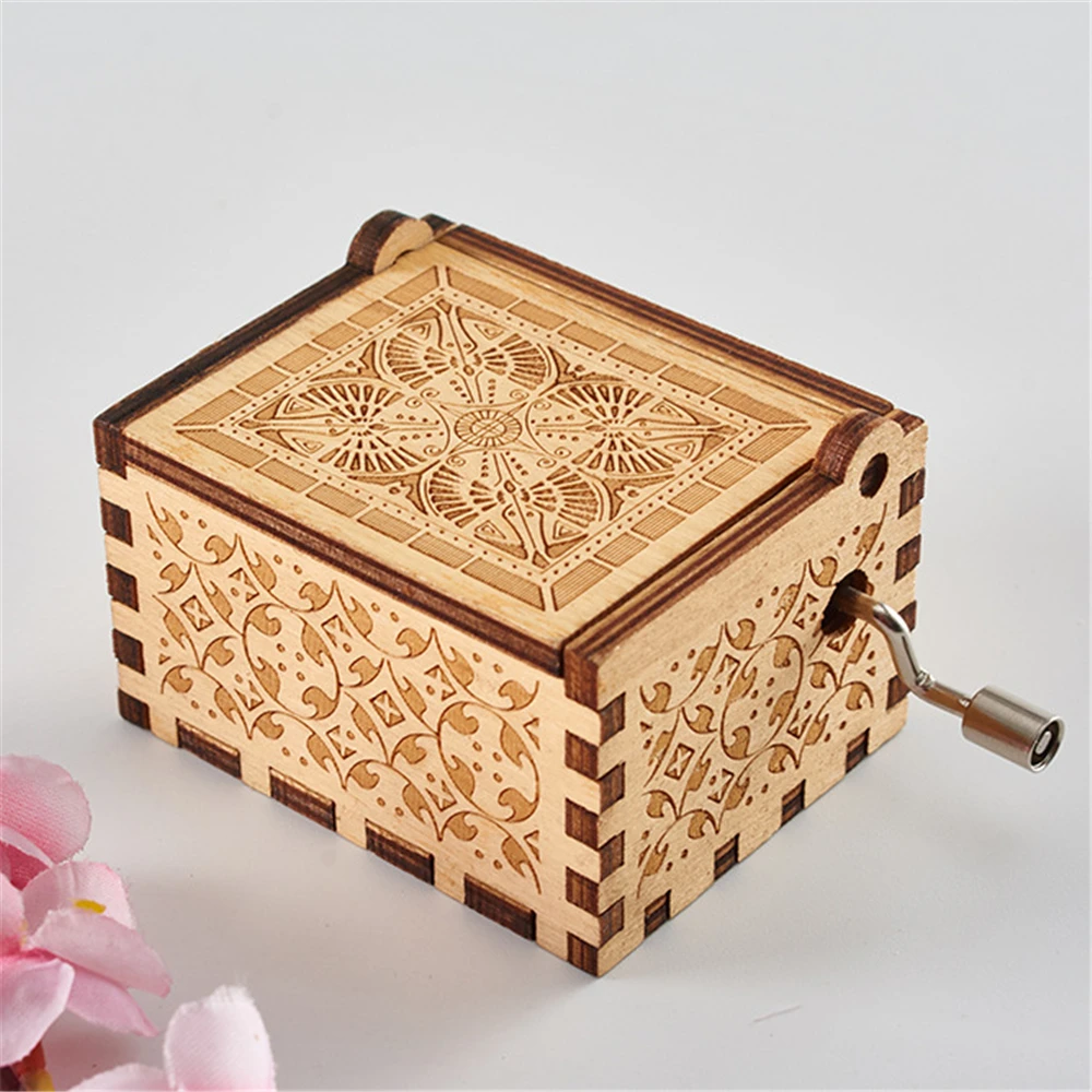Product: Wooden Music Box

Description: A small, ornate, handmade music box with intricate floral carvings on the sides and top. This hand-cranked wooden music box features a metal crank handle on one side, perfect for playing classic tunes. Pink flowers are partially visible in the foreground.
