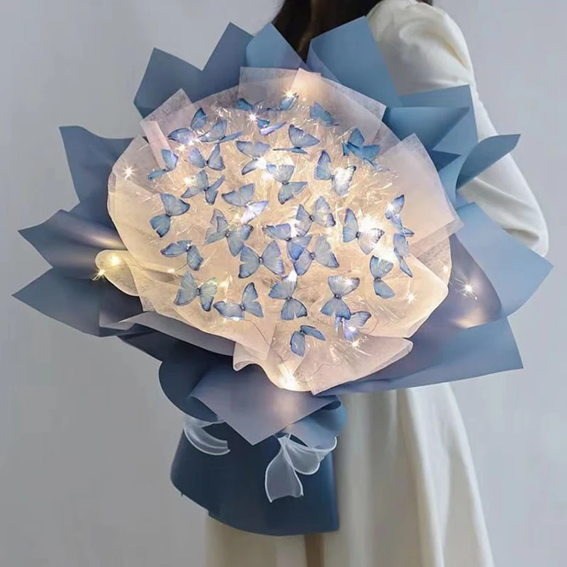 A person holds the Creative Handmade Butterfly Bouquet, featuring charming blue butterfly shapes and artificial flowers wrapped in elegant blue paper. Tiny white lights delicately sprinkle the arrangement, harmonizing beautifully with the translucent material—perfect for home decoration.