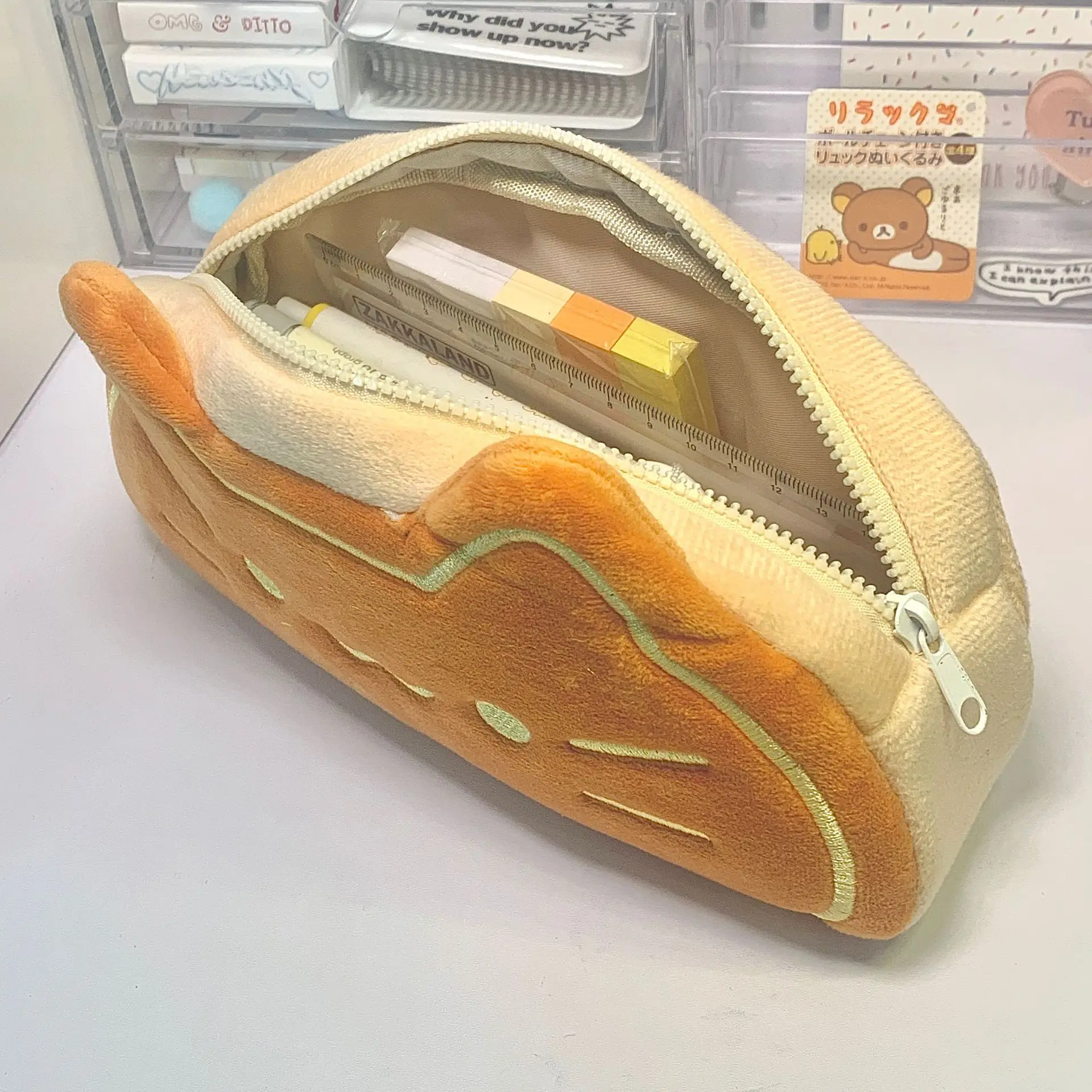 The KittyNook Cat Co. Caramel Cat Plush Pencil Case is unzipped, showing plush school supplies on a light desk.