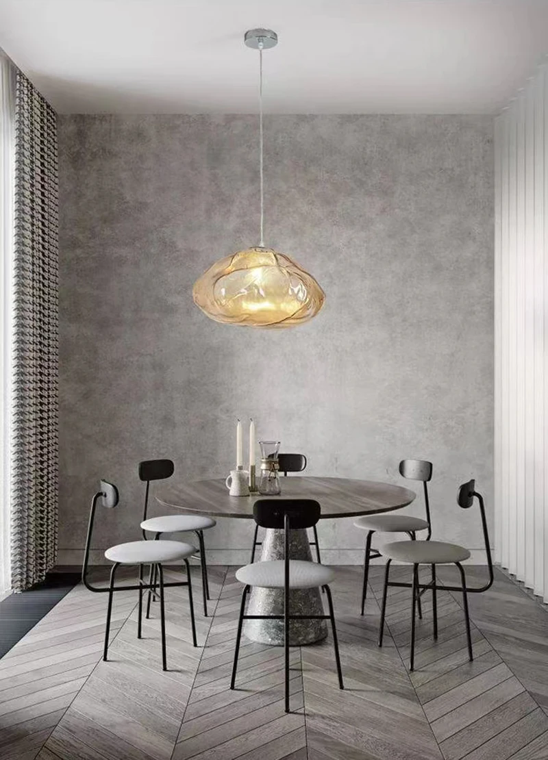 A modern dining area features a round table, four black chairs, two candles, and the Luxe Cloud Glass Pendant Light, set against grey textured walls and chevron-patterned flooring.
