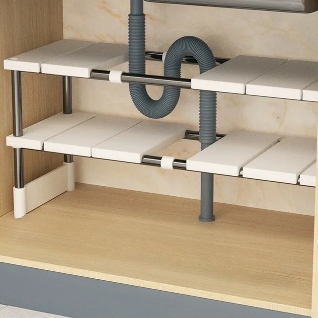An expandable under sink organizer made of stainless steel and white panels, adjusted to fit neatly around a sink's drain pipe.