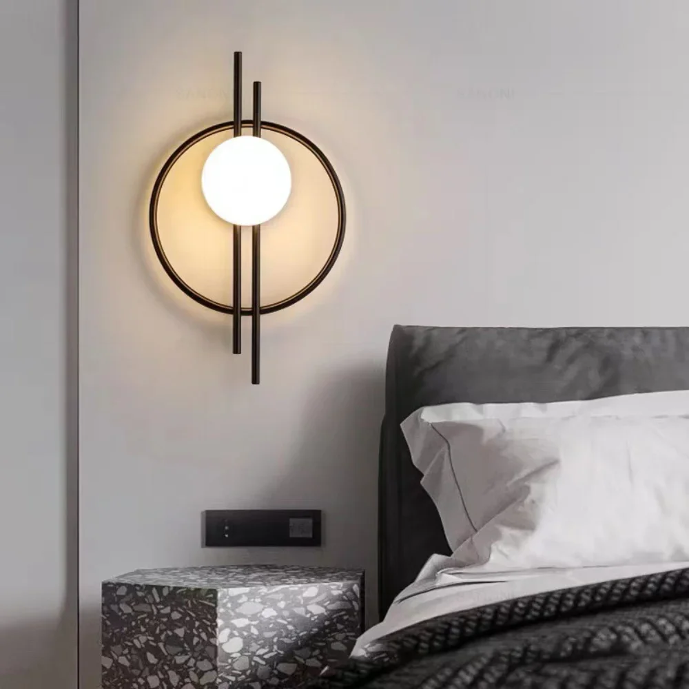 A Circular Wall Sconce with Dual Vertical Rods, featuring a glowing white sphere and a modern circular design, is mounted on a white wall next to a bed adorned with gray linens and accompanied by a terrazzo nightstand.