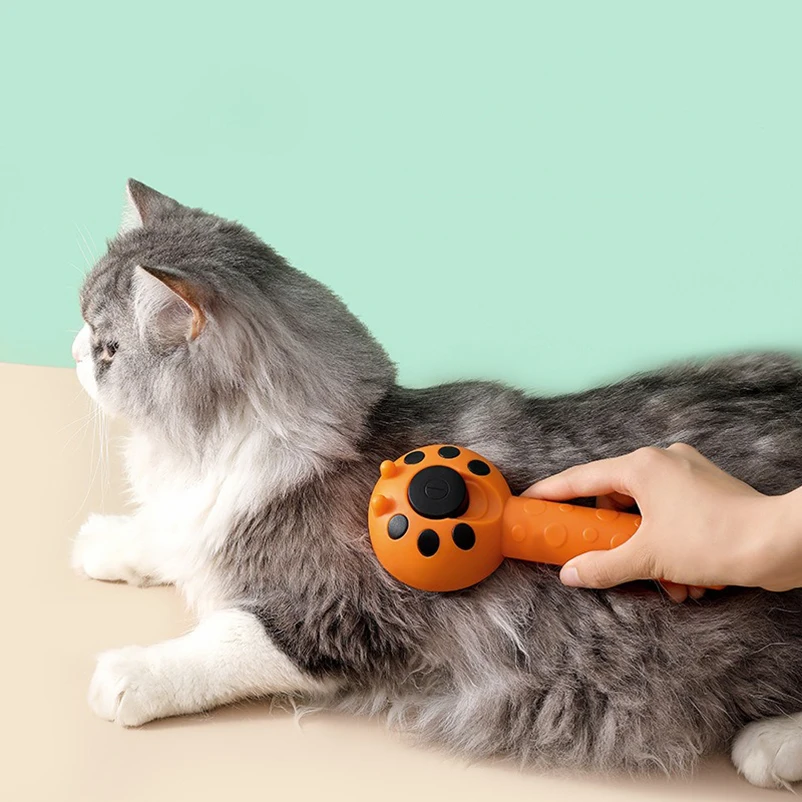 Chat gris et blanc allongé sur une surface beige, en train d'être brossé avec une brosse autonettoyante orange. Toilettage animaux, accessoires et brosses pour chats.