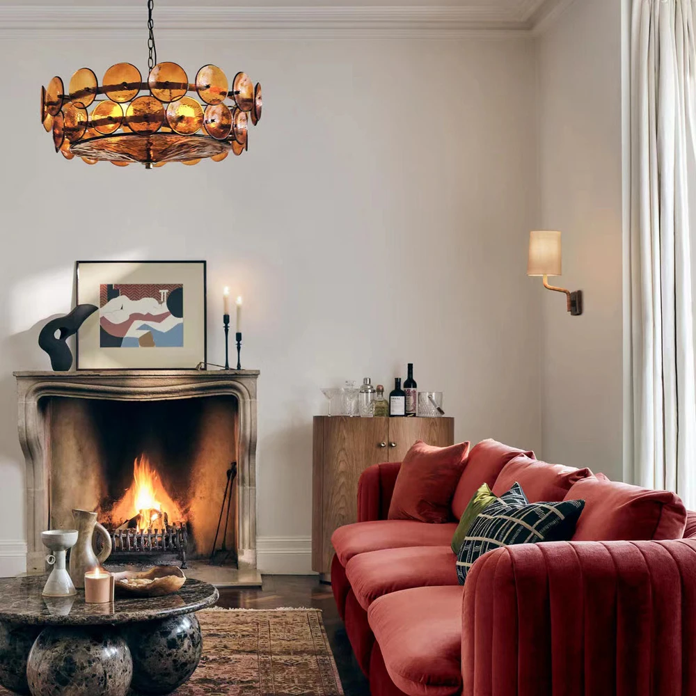 A living room with a red velvet sofa, lit fireplace, round coffee table, wall art, and an Italian Retro Pendant Light. A wooden cabinet with wine bottles stands in the corner near curtained windows.