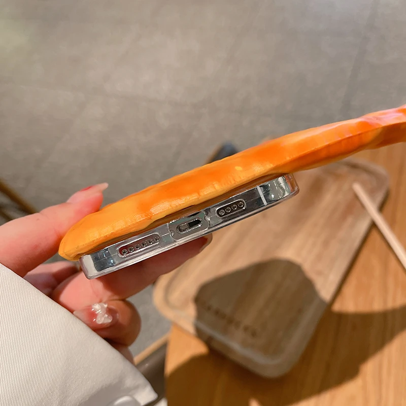 A person holds a smartphone featuring the Chicken Drumstick iPhone Case, showcasing its bright orange and unique design that accentuates the bottom part of the device, including the charging port and speakers, against a wooden table background.