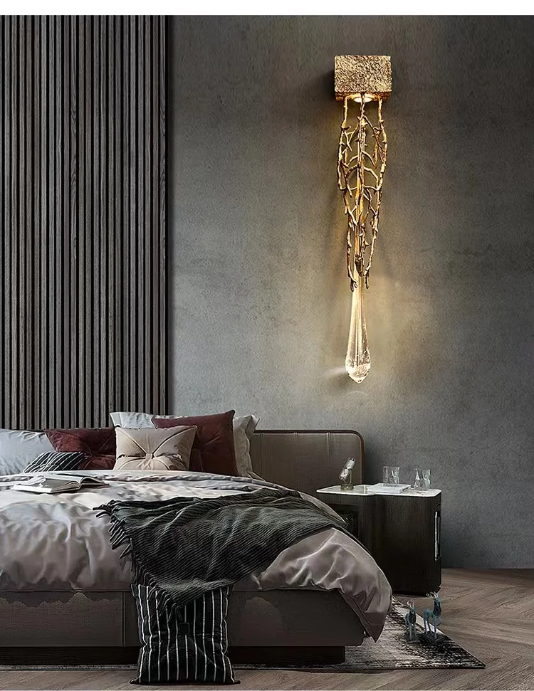 In the elegantly designed bedroom, the Luxury Vertical Wall Light showcases intricate metalwork and a teardrop crystal accent above a contemporary bed adorned with plush pillows and cushions. A side table holds glasses and water, all beautifully illuminated by this luxury wall sconce.