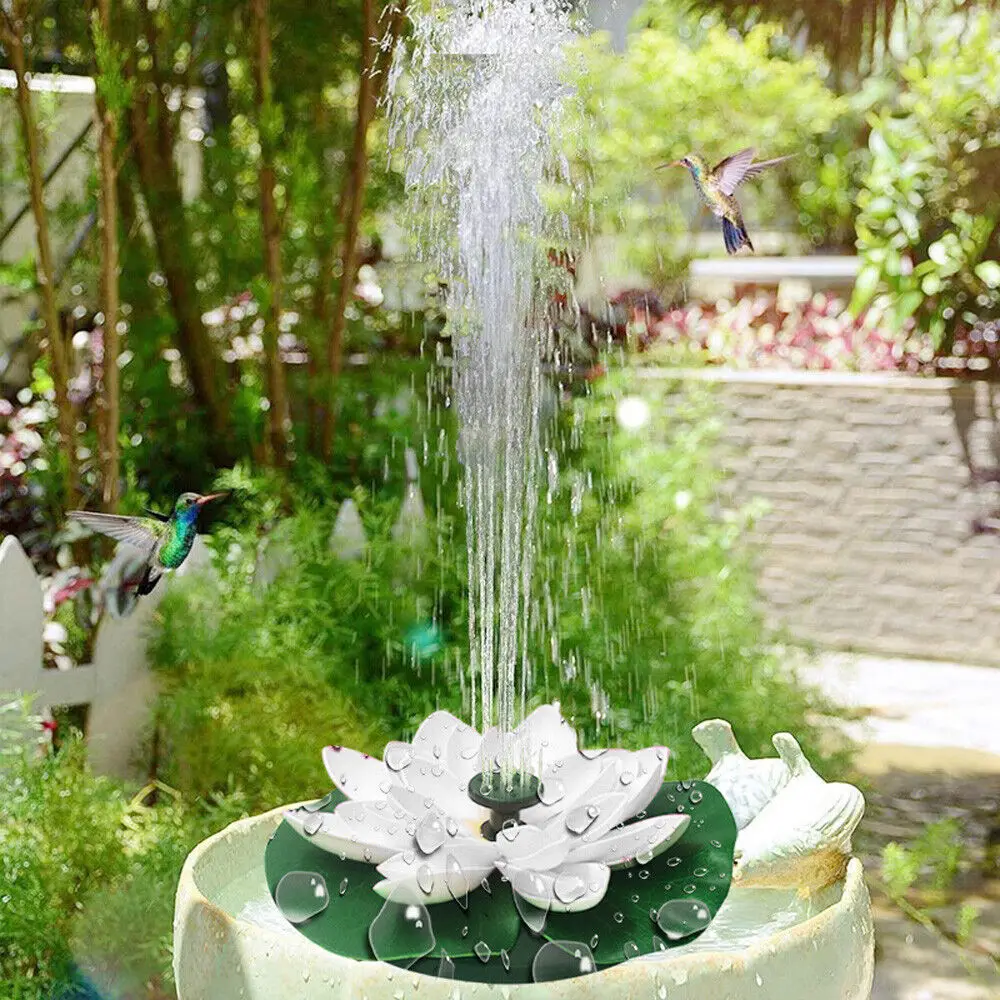 A lotus-shaped solar fountain with water spraying upward sits in a garden pond. Two hummingbirds are flying nearby. Green foliage and plants surround the area on a sunny day, enhanced by the Lotus-shaped Solar Fountain's floating design.