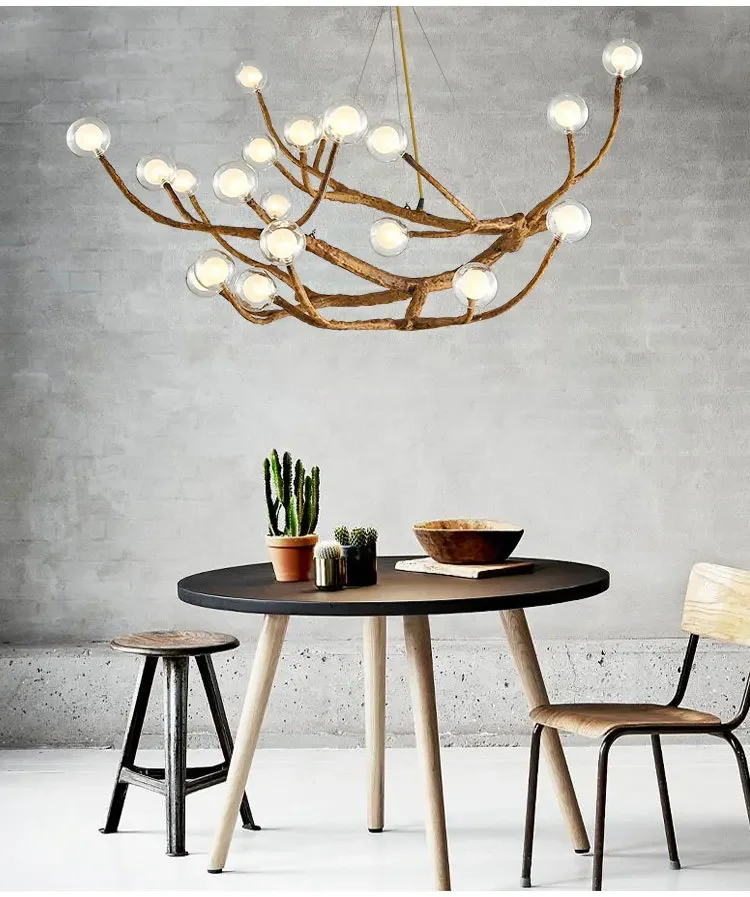 A modern dining area features a round table, two chairs, and a stool. A cactus and wooden bowl sit on the table. Above, the Branch Pendant Light with glass orb shades provides an organic modern style against the neutral-toned wall background.