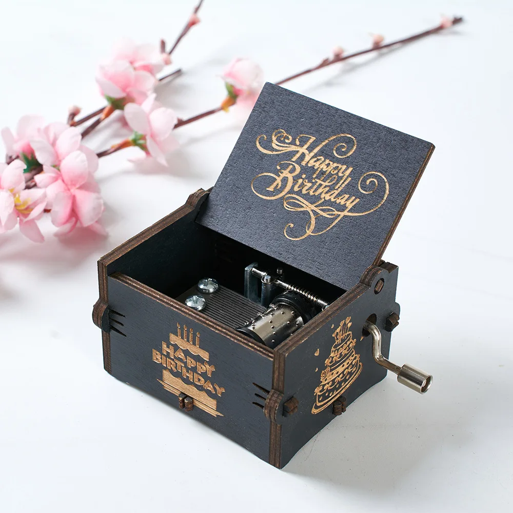A Happy Birthday Music Box, featuring a sleek black design and an open hand crank mechanism with gold-colored engraving, is beautifully positioned near pink flowers. This elegant wooden music box is an ideal birthday gift that combines beauty and sentiment in one charming package.