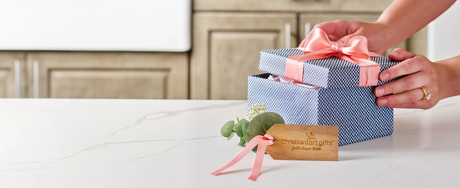Woman packing blue gift box with pink ribbon on kitchen counter Christian Art Gifts tag
