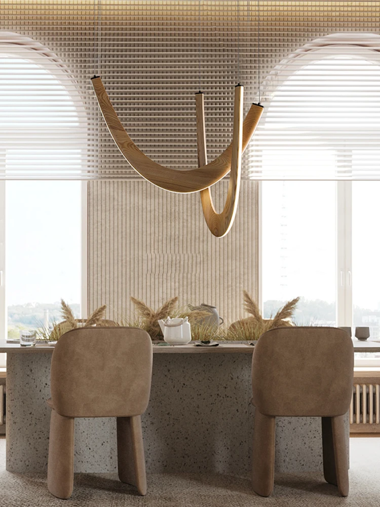 A modern dining area features two beige chairs, a stone table, dried plant decor, and a Double Curve Wood Pendant Light above, all set in front of large windows with horizontal blinds.