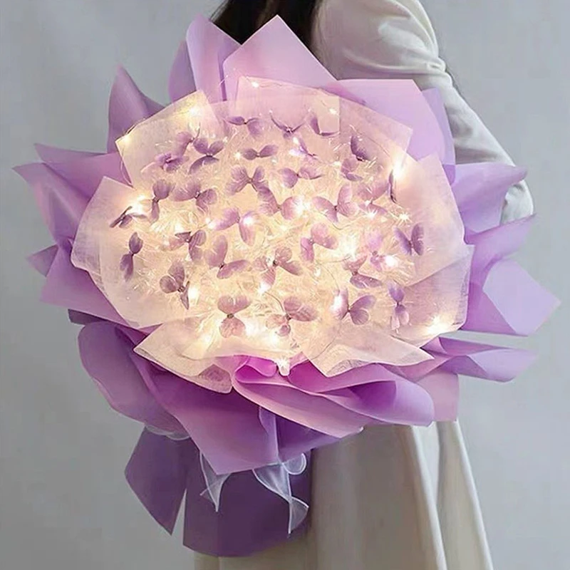 A person is holding a Creative Handmade Butterfly Bouquet, featuring purple and white flowers adorned with soft glowing lights, elegantly wrapped in sheer paper. This exquisite arrangement of artificial flowers makes for a captivating home decoration. The person is dressed in a light-colored coat.
