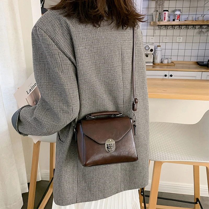 Pruuni värvi väike crossbody kott sangaga, seljas naisel, kes kannab halli ruudulist bleiserit köögi taustal.