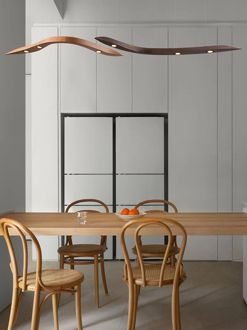 Minimalist kitchen with a light wood dining table, four matching chairs, a glass of water, a bowl of oranges, and two Spiral Wood Pendant Lights overhead for an organic modern lighting touch.